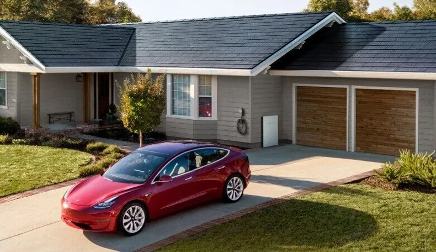 Tesla-Solar-Glass-Roof-V3_2 A red car is parked in a driveway in front of a single-story house with solar panels on the roof. The garage has wooden doors, and the lawn is neatly maintained.