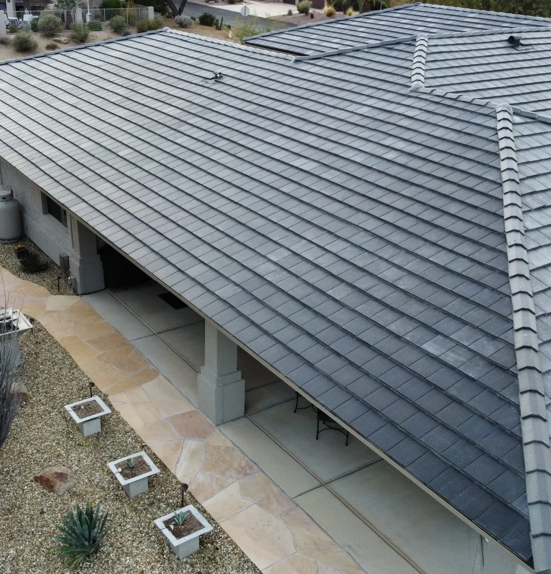 c14c29_ba3adf944adc412cb16c62bcaa16e4de~mv2 (1) Aerial view of a house with a gray tiled roof, a covered patio, stone pathways, and landscaped yard with plants and mulch.
