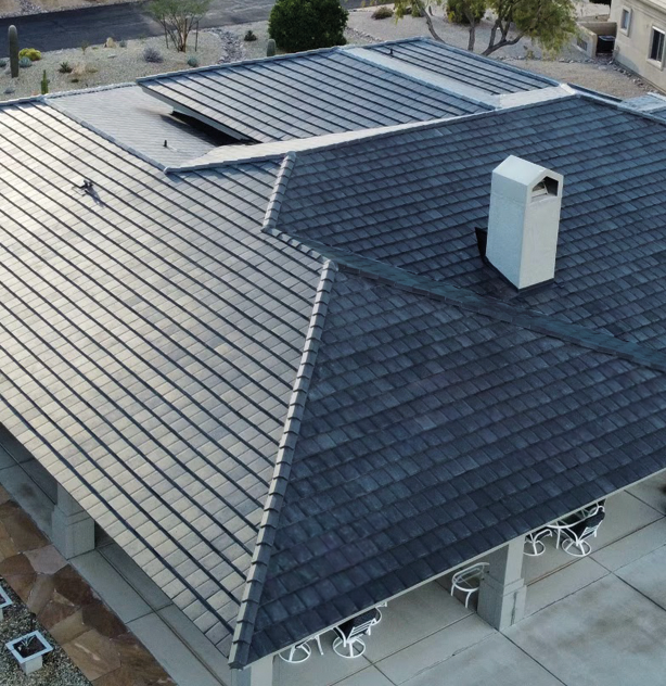 c14c29_c7f2786b491b46e98b456ce0db71a6a4~mv2 Aerial view of a house with a dark gray, shingled roof, featuring a skylight and a chimney. The surrounding area includes a landscaped yard with desert plants and outdoor seating.