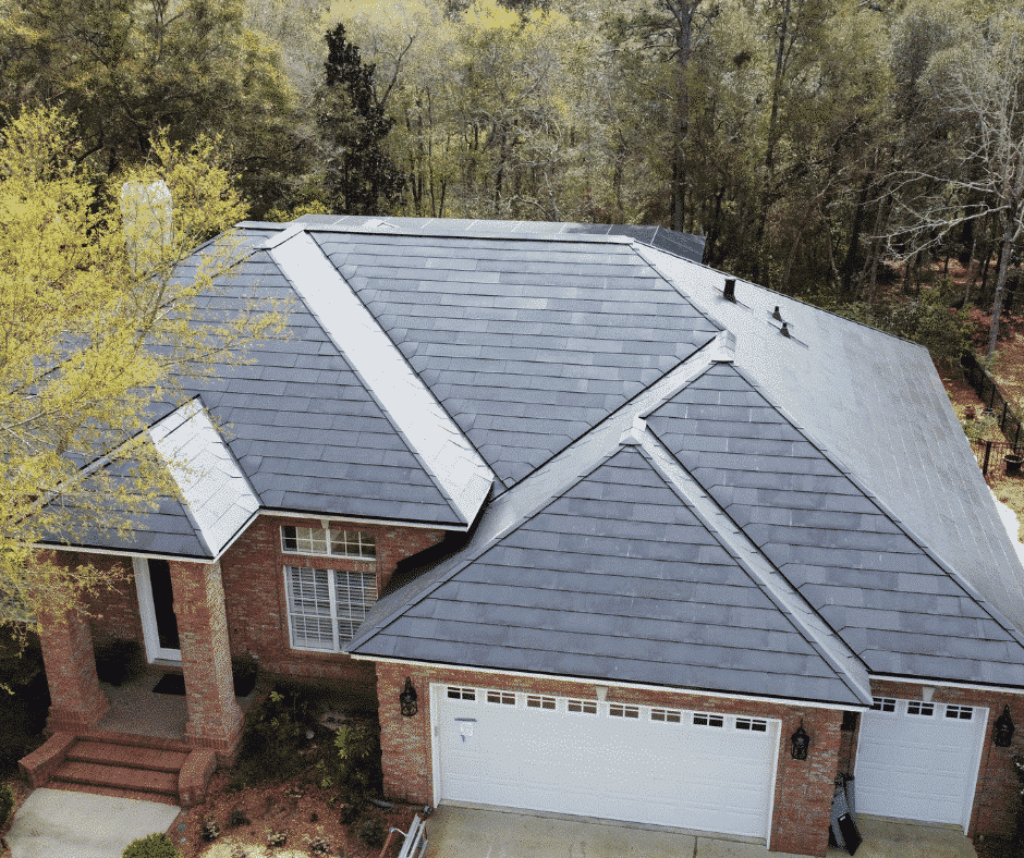 grey telsa roof Aerial view of a house with a sleek, dark-colored metal roof surrounded by trees. The house features a three-car garage and a brick exterior.