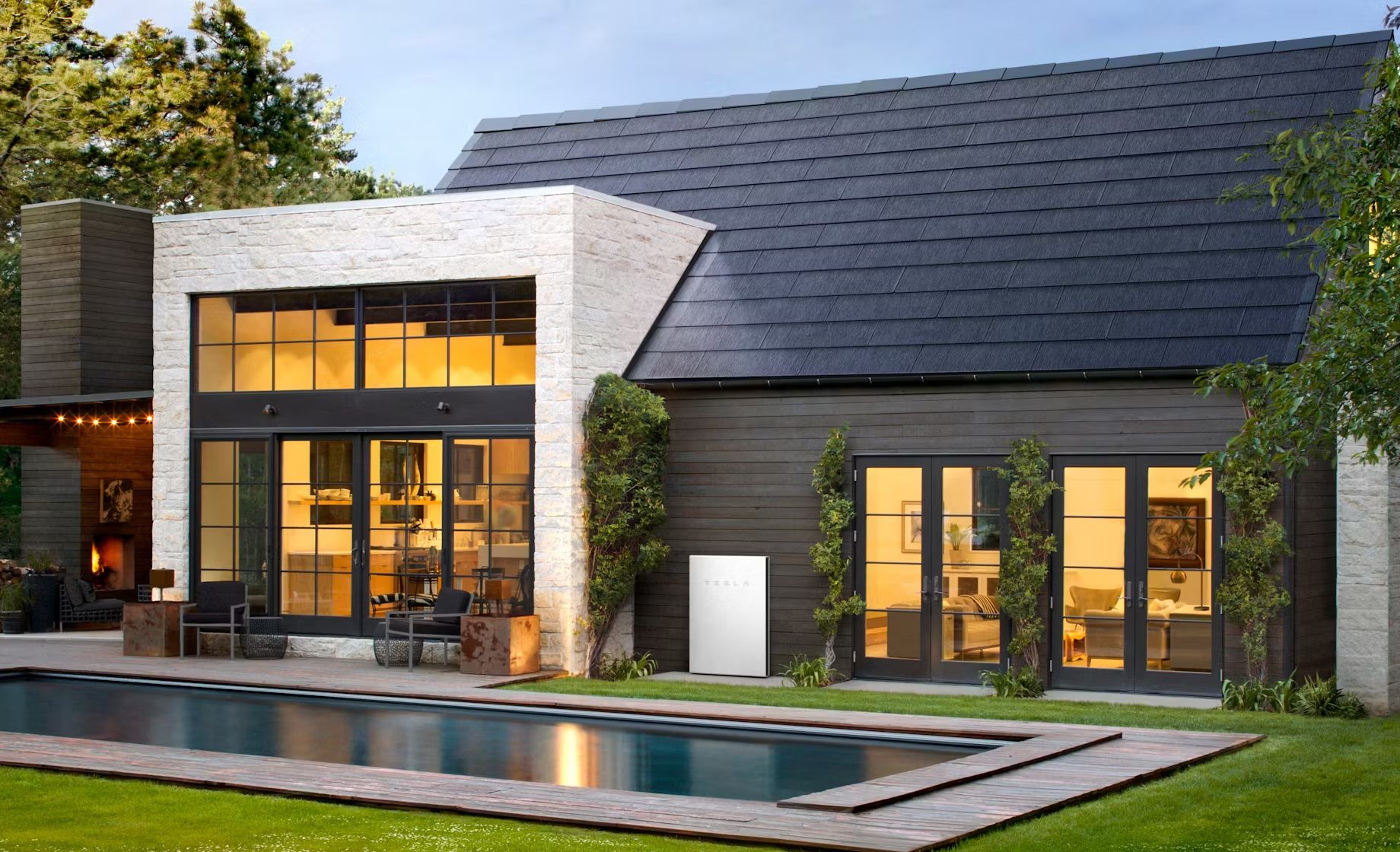 tesla roof home copy Modern house with large windows, solar roof tiles, and a rectangular pool in the foreground. Lawn surrounds the pool, and the house features multiple stories with evening lights on inside.